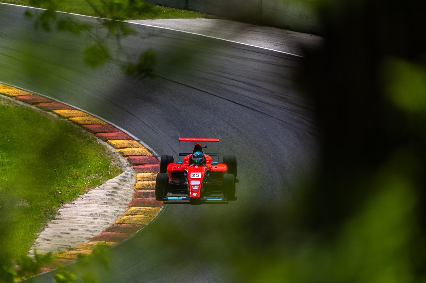 The red No. 25 Ligier JS F4 car turning into a corner at Road America, seen through roadside foliage.
